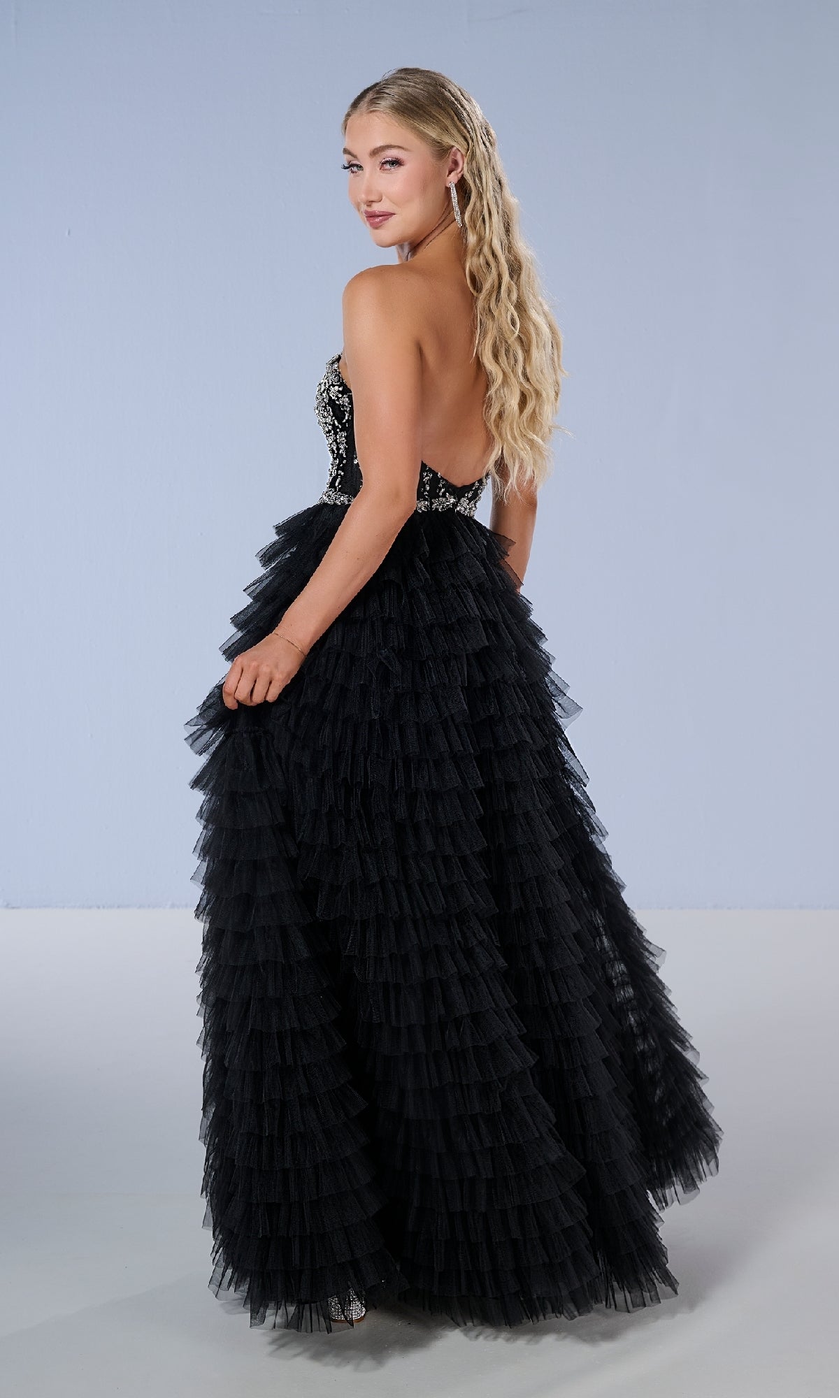 A woman with long blonde hair poses against a plain light background, looking over her shoulder in the Tiffany 16220 long prom dress, which features a strapless bodice and a tiered ruffle skirt.